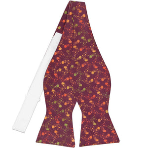 A self-tie bow tie, untied, with colorful leaves on a dark burgundy background