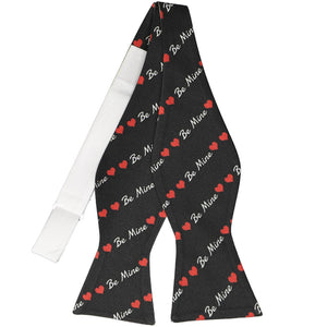 An untied self-tie bow tie in black, with a heart and be mine striped pattern