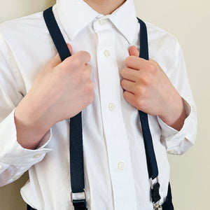 A boy holding onto the straps on a pair of navy blue suspenders with a white dress shirt
