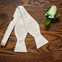 Load image into Gallery viewer, An untied ivory floral self-tie bow tie, displayed on a wood floor with a white rose boutonniere