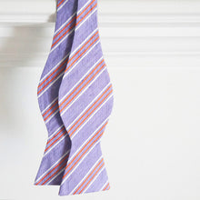 Load image into Gallery viewer, A purple and orange striped self tie bow tie, untied, handing from a white wood trim background