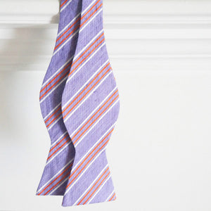 A purple and orange striped self tie bow tie, untied, handing from a white wood trim background